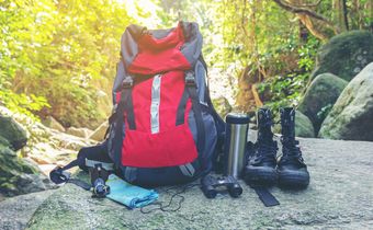 Auf dem Foto sieht man einen Rucksack, ein Fernglas, Wanderschuhe, Trinkflasche, Kompss und ein Handtuch, die auf einem Felsen stehen.