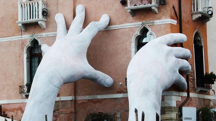 Auf dem Bild sieht man eine Skulptur von Lorenzo Quinn: Zwei riesige Hände, die ein Haus "stützen"