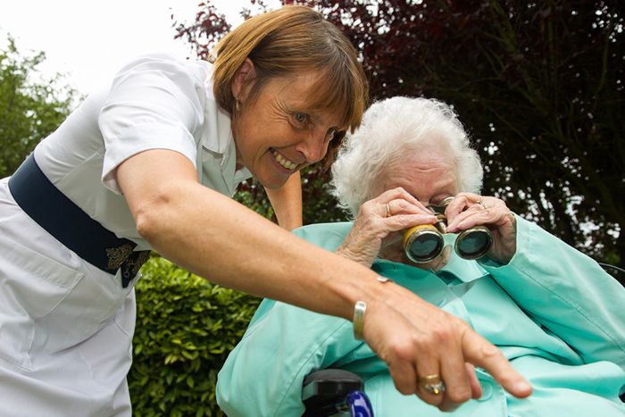 Auf dem Foto sieht man eine alte Frau im Rollstuhl, die durch ein Fernglas schaut. An ihrer Seite ist eine etwas jüngere Frau.